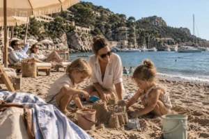 Nanny im Familienurlaub betreut zwei Kinder beim Sandburgbauen am Strand, während die Eltern entspannt im Hintergrund sitzen.